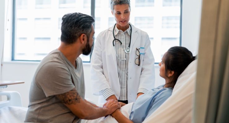 Image of a doctor speaking to a patient in a hospital bed with her spouse sitting nearby 